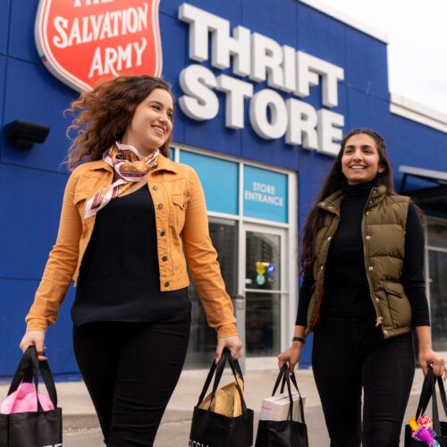 Two young women holding shopping bags after a trip to their local Salvation Army Thrift Store with the store pictured in the background. Shift to thrift to save money, save the planet and help neighbours in need in your community