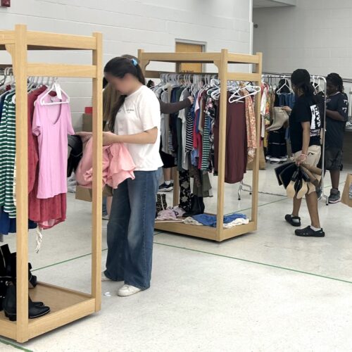 clothing racks with different t-shirts, sweaters, jeans, and longsleeves with young females choosing items square crop