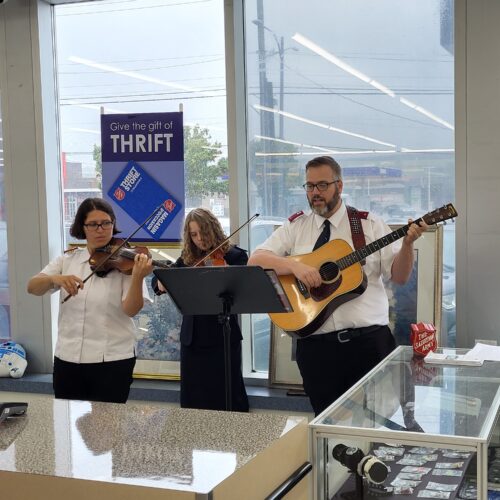 A guitarist singing, and 2 violinists standing performing inside a thrift store with a music stand square